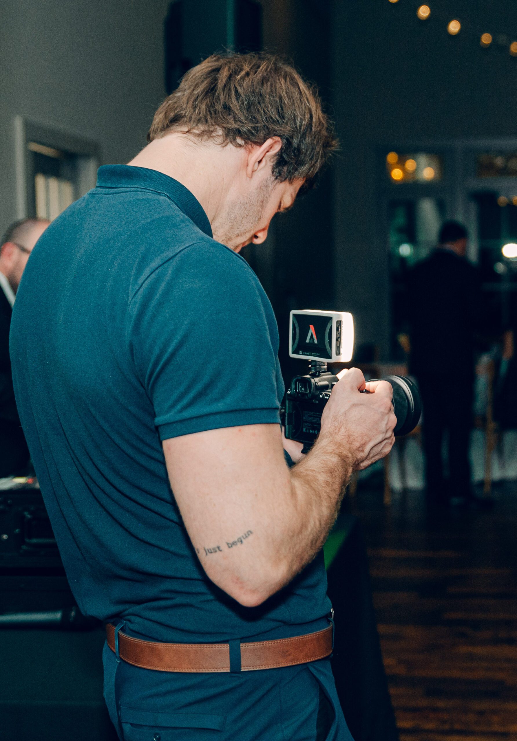 A person with short hair wearing a blue polo shirt and brown belt is holding a camera and looking through the viewfinder. They have a tattoo on their forearm. The setting appears to be indoors with dim lighting and blurred people in the background.