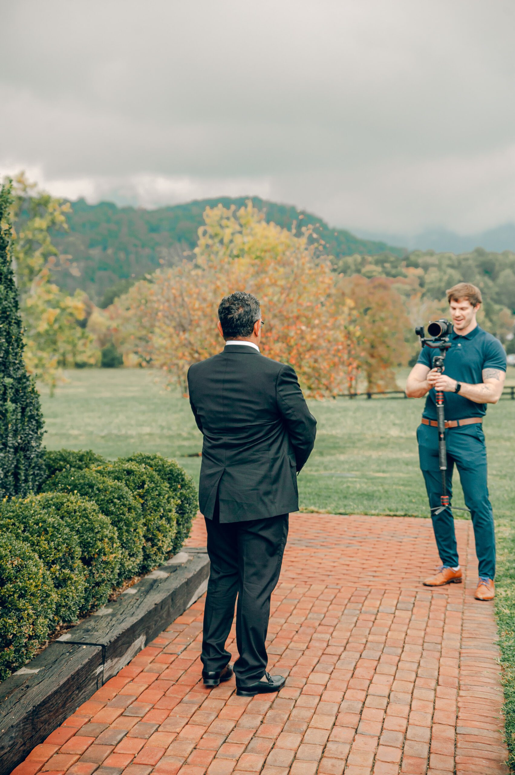 A man in a black suit with his back to the camera stands on a brick path, facing a man operating a camera on a gimbal. They are outdoors, with a lush green lawn, bushes, and a scenic backdrop of hills and cloudy skies.