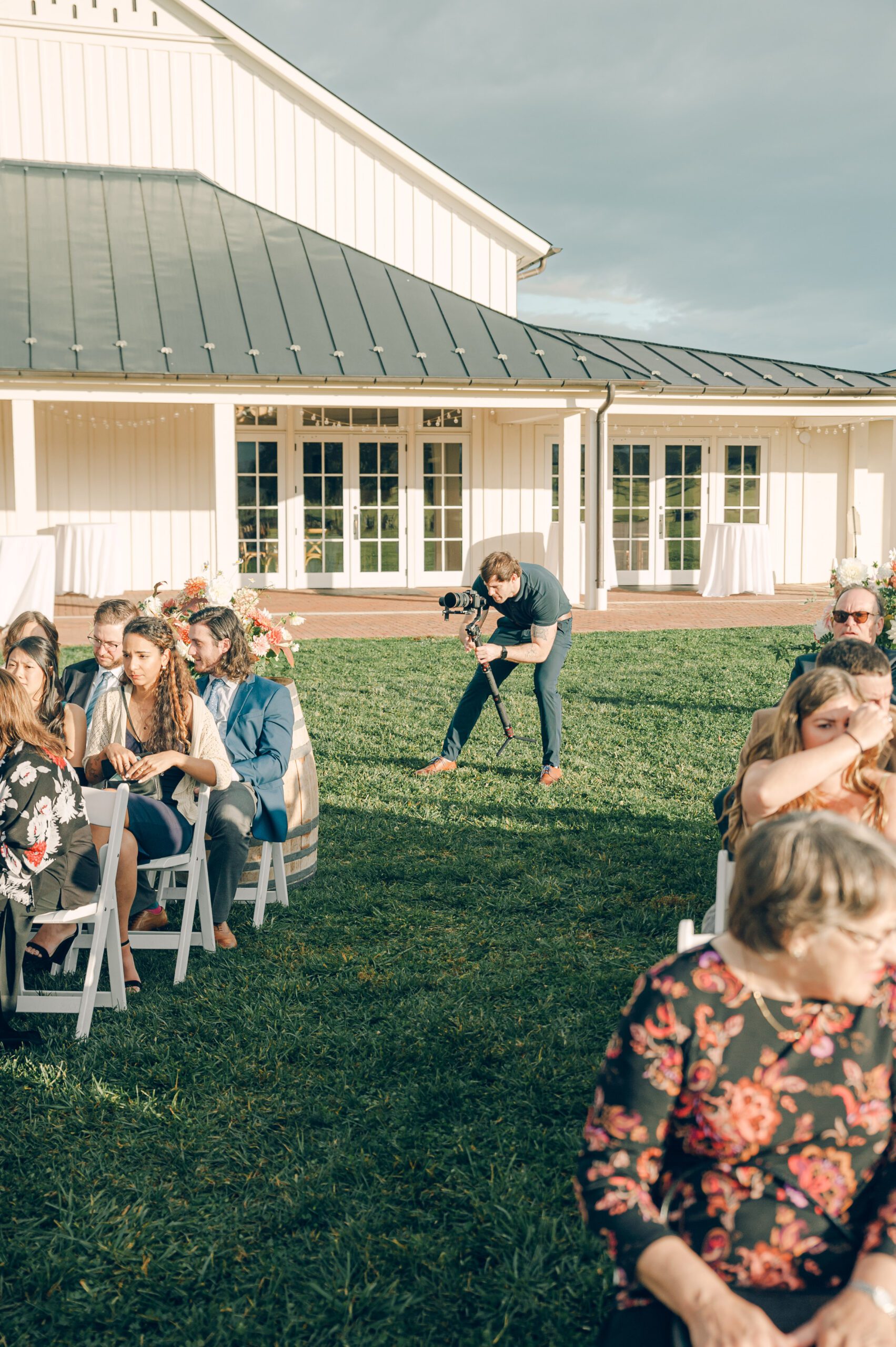 A photographer crouches with a camera aiming at a group of people seated on white folding chairs on a lawn during an outdoor event. A white building with large windows and a dark roof is in the background. Some people chat, and greenery surrounds the area.