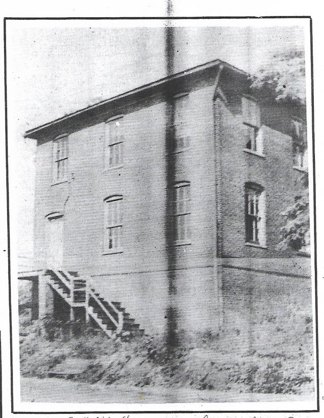 A black-and-white photograph of an old two-story brick building. The building features a staircase leading to an elevated entrance on the left side. There are several windows, with foliage partially visible on the right. The image shows vertical lines likely due to damage.