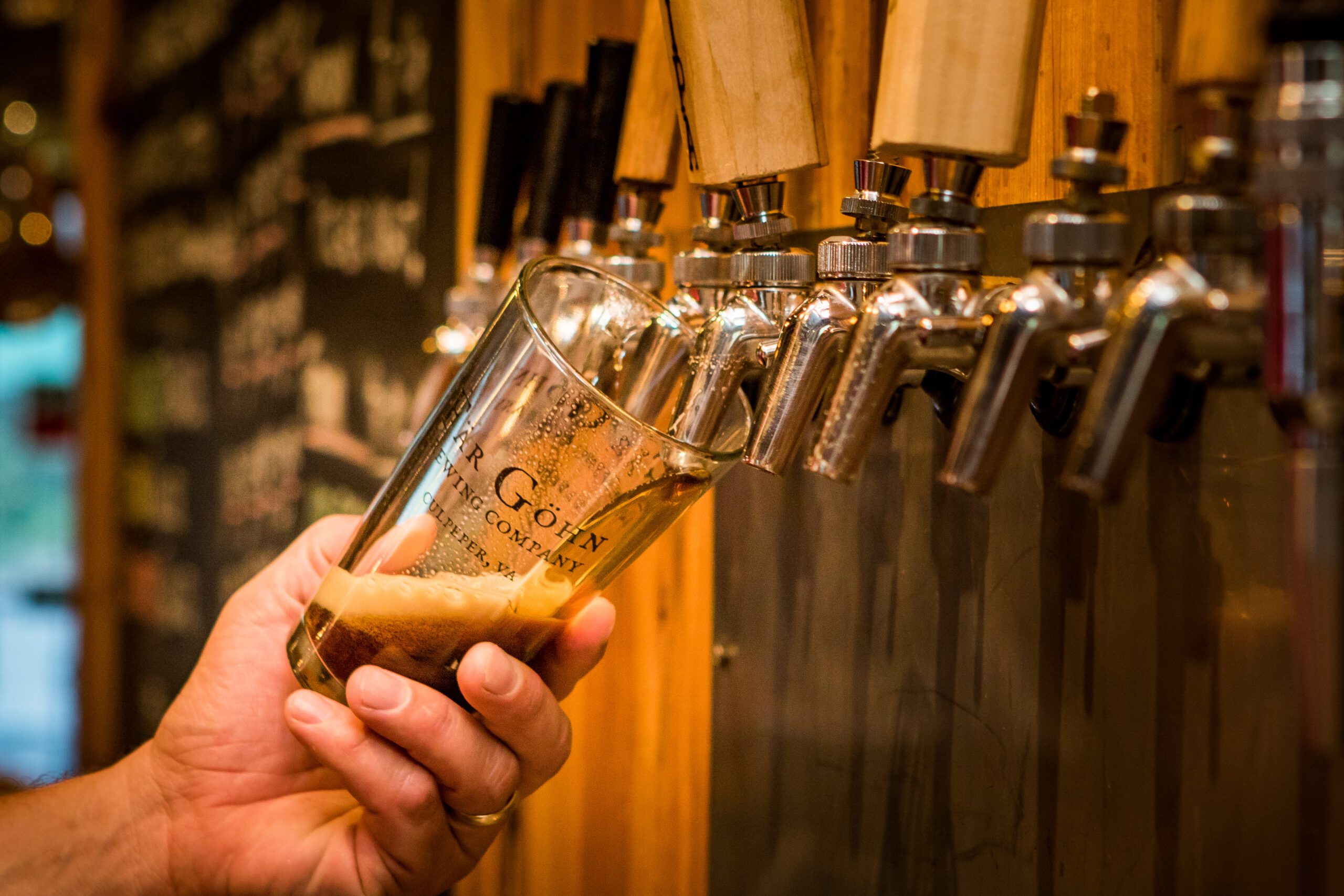A hand holding a glass tilted at an angle under a beer tap, pouring a dark beer with foam into the glass. The background features multiple beer taps and a chalkboard with blurred text, hinting at the next stop on your beverage trail in this cozy bar setting.