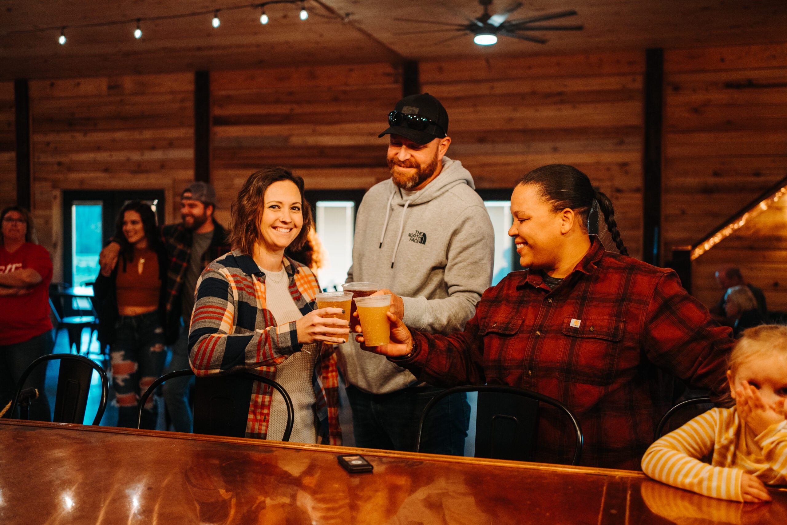 A group of people gathering indoors, with two of them at the bar holding and clinking glasses while smiling. The background features wood-paneled walls and several people interacting in a relaxed setting, as if they've just completed a local beverage trail.