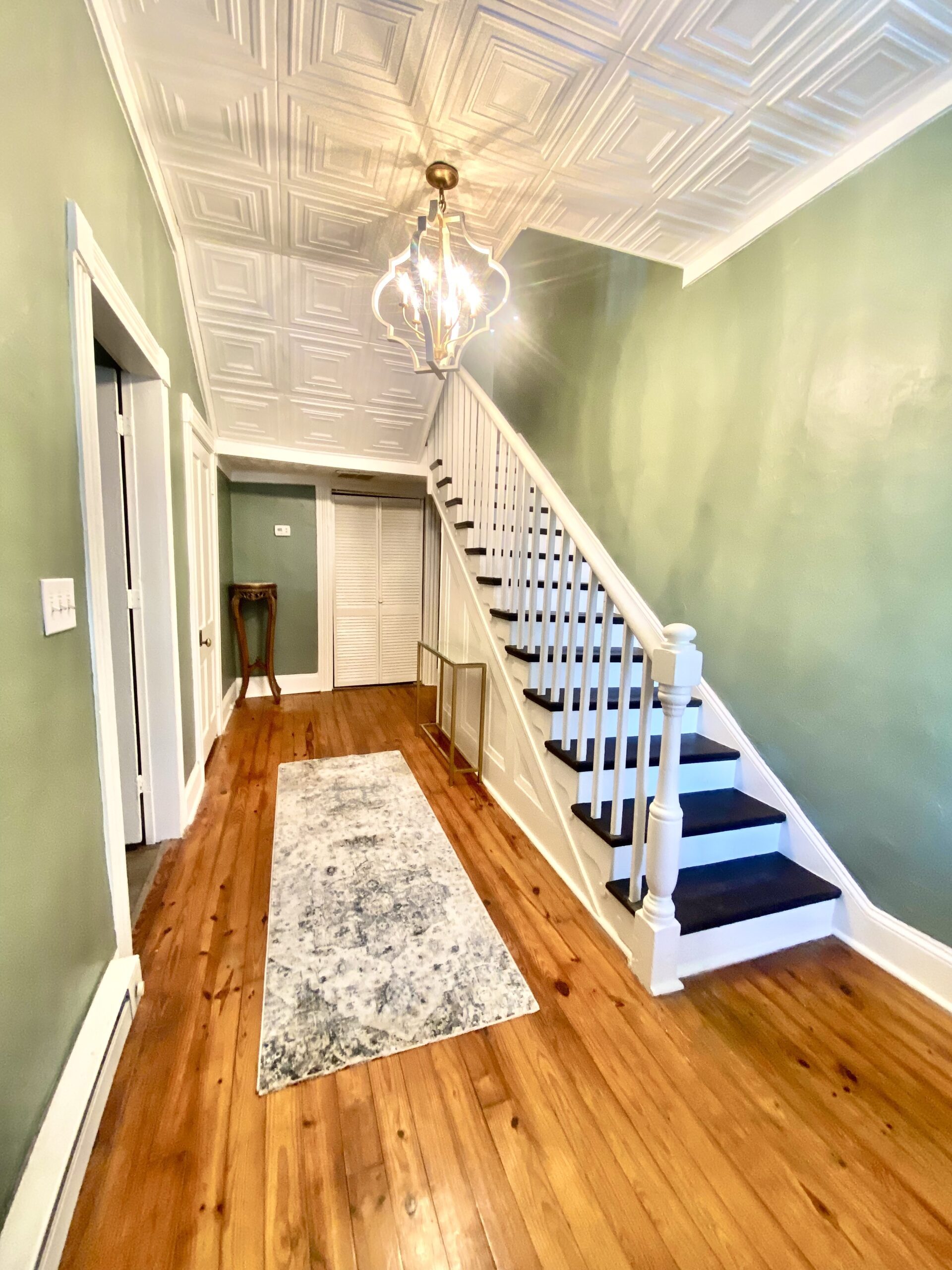 A well-lit hallway with light green walls and hardwood floors in Hill House features a white staircase with dark treads. A patterned runner rug lies on the floor, and a small table is placed against the wall near the closed double doors at the far end. A chandelier hangs from the ceiling.