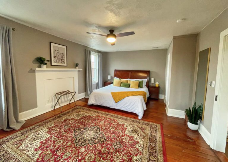 A cozy bedroom, perfect for a spring escape, features a large bed with white bedding, yellow pillows, and a wooden headboard. A ceiling fan is above the bed, and gray curtains cover the windows. A decorative rug, potted plant, and fireplace add charming touches to the room.
