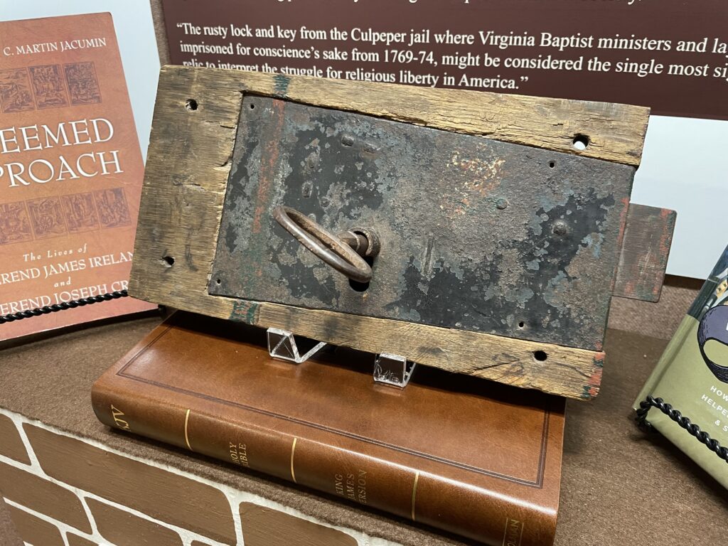 A close-up of an old, rusted lock and key mounted on a wooden board displayed in the museum exhibit. The lock is weathered, indicating age. The display is set against a backdrop with historical text and books related to Baptist ministers and religious liberty in America.