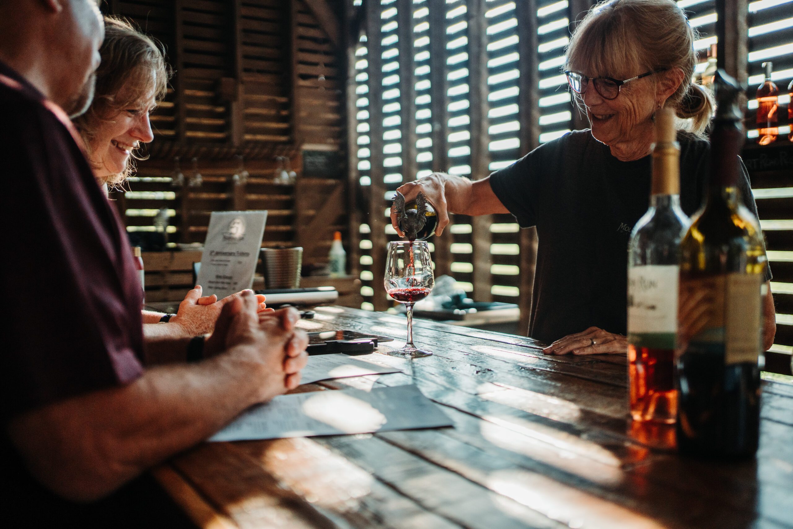 A woman with glasses and a ponytail pours red wine from a bottle into a glass on a wooden table, creating a delicate beverage trail. Two people seated at the table, one man and one woman, smile and watch as sunlight filters through wooden slats in the background, casting a warm glow.