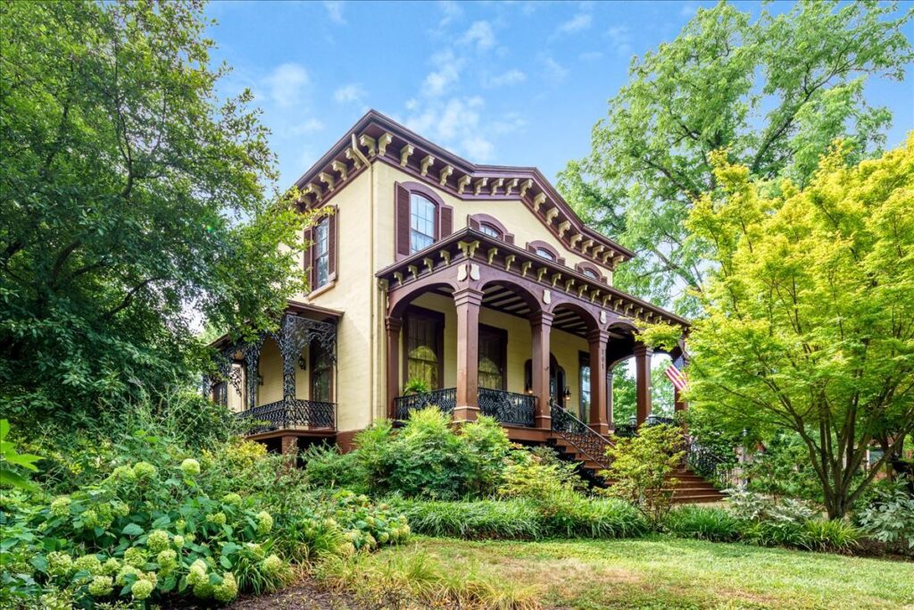 A Victorian-style Hill Mansion with a wraparound porch featuring intricate railings, surrounded by lush greenery and trees. The house has large windows with shutters and detailed wooden trim under the roof. The sky is blue with scattered clouds.