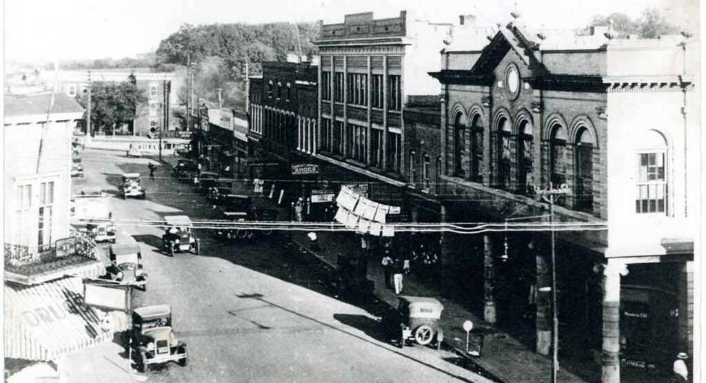 A historic black and white photo shows a busy street lined with early 20th-century buildings and cars.