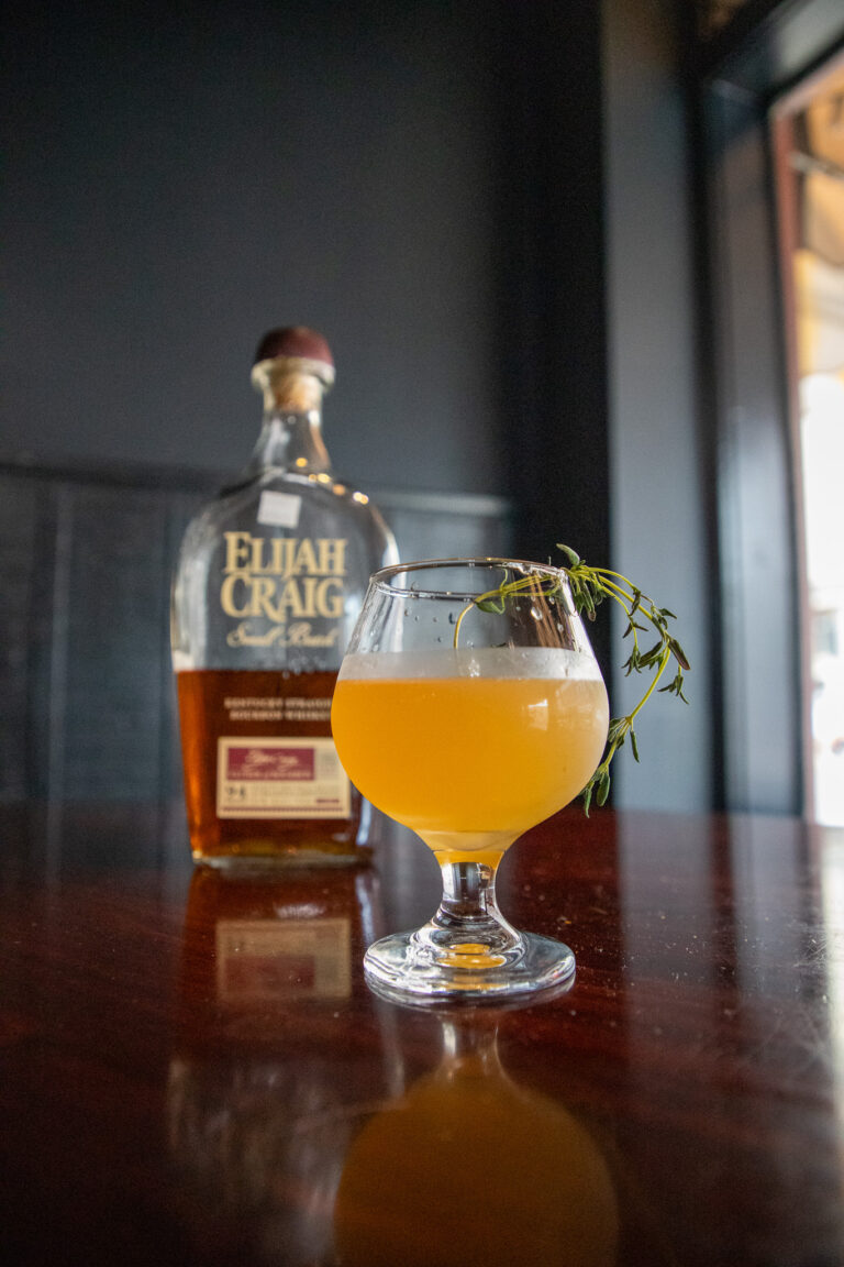 bottle of Elijah Craig and a yellow drink with a green sprig coming out